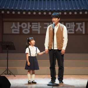 학교 축제 무대에서 마이크를 들고 노래하는 아버지와 손을 흔드는 딸의 모습